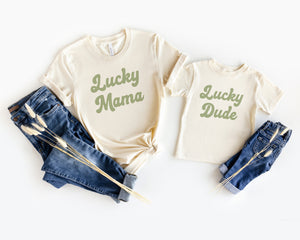 Adult natural shirt next to matching toddler shirt with lucky mama and lucky babe print in green retro script text, laying flat on white background styled with blue jeans